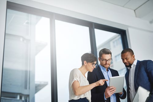 Three professionals collaborate, discussing something on a tablet in a bright, modern office. They appear focused and engaged in teamwork.