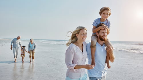 A family enjoys a sunny day on the beach. A man carries a smiling child on his shoulders, walking alongside a woman. An elderly couple and child stroll behind them.