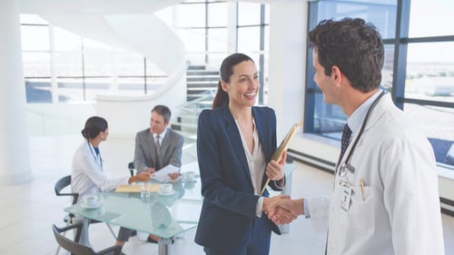 A doctor shakes hands with a smiling businesswoman holding a file in a bright office. In the background, two people converse at a glass table.
