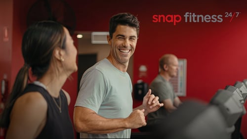 Two happy Snap Fitness members run on a treadmill inside a 24/7 gym.
