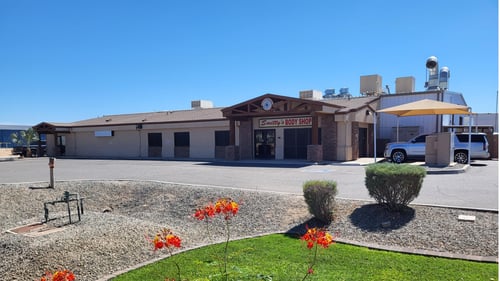 The attached image shows the exterior of “Smitty’s Body Shop,” a one-story building with a beige stucco facade and brown brick accents around the entrance. The entrance is covered by a large wooden gable with a sign that reads “Smitty’s BODY SHOP” in red and black letters. The building is surrounded by a paved parking area, with a few small bushes and a patch of landscaped gravel with bright red and yellow flowers in the foreground. To the right of the main building, there is a covered area with a beige canopy, under which a white SUV is parked. The sky is clear and blue, and the overall setting is neat and well-maintained, suggesting a professional and welcoming business environment.