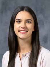 Jenna is smiling in front of a gray background with a pink blouse.
