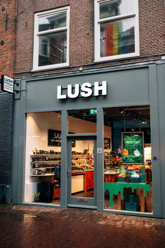 Exterior view of the Haarlem shop. Central doorway with glass windows either side framed in black. Large Lush symbol above the door. Products displayed in the right side window, interior of the shop can be seen from the outside.