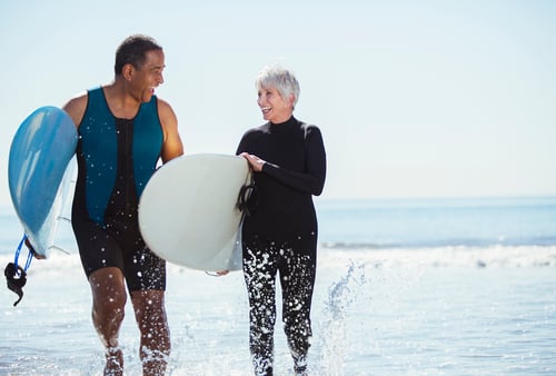 A man and woman, both in wetsuits, joyfully walk through shallow ocean water, holding surfboards and smiling, with a bright blue sky overhead.