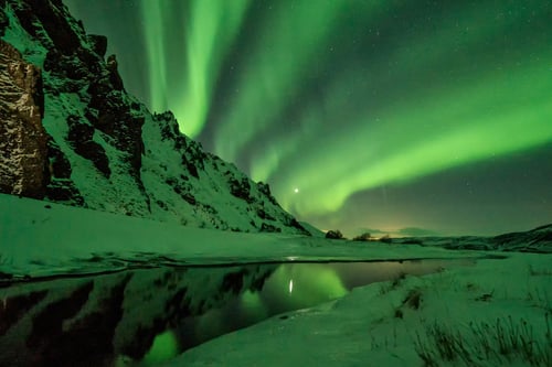 Aurora borealis reflecting over frozen lake with snow-covered mountain cliff at night.