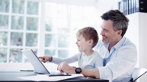 Father and son smiling at a laptop in a bright room with large windows and natural light. The scene conveys happiness and bonding.