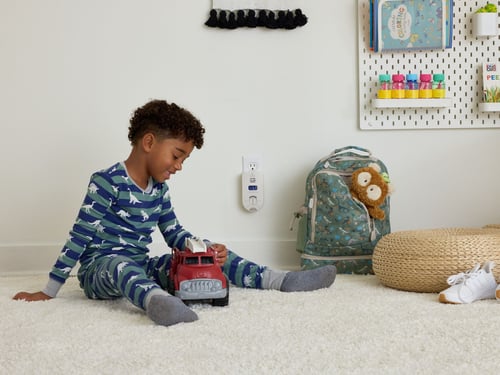 Child sitting on the floor playing with a fire truck. Near him are a backpack and shoes. Outlet on the wall with a carbon monoxide alarm.