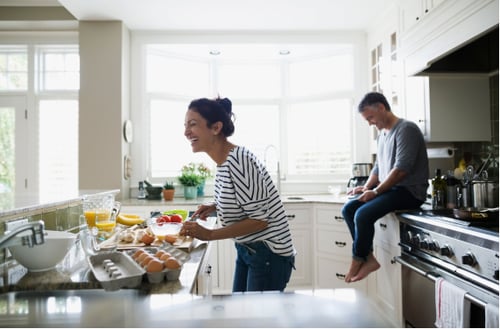 A woman in a striped shirt is joyfully preparing breakfast in a bright kitchen. Nearby, a man sits on the counter, smiling and relaxed. Egg cartons, fruit, and juice are on the island. Natural light streams through large windows, creating a warm, cheerful atmosphere.