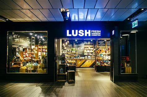 Exterior view of the shopfront. Large open doorway with illuminated Lush sign above. Glass window to left side of door with product display.