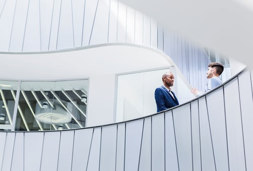 Two people engage in conversation on a modern, curved balcony with white and gray walls in an office setting, conveying a professional tone.