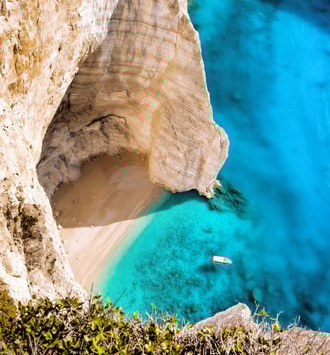 High-angle view of a secluded sandy beach in a cove, bordered by tall white cliffs and bright turquoise water with a small boat floating nearby.