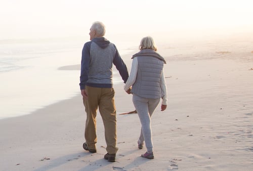 Two older adults with gray hair holding hands while walking together on a sandy beach at sunset or sunrise. The person on the left wears a blue hoodie and tan pants, while the person on the right wears a gray puffy vest and leggings. They are viewed from behind as they walk toward the calm ocean, with soft golden light illuminating the beach and peaceful waters in the background.