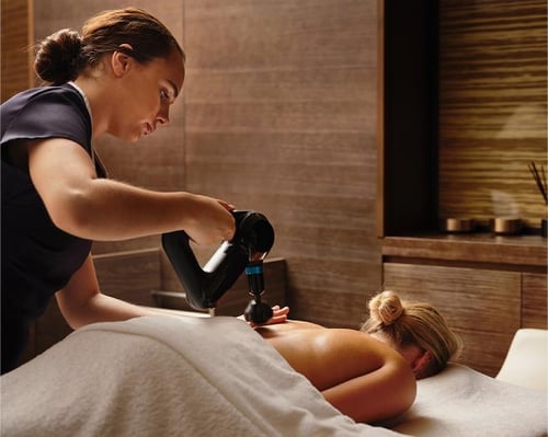 Massage therapist using a handheld massage device on a client's back in a serene spa setting.