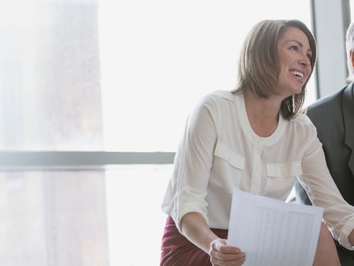 A woman in a white blouse and red skirt smiles brightly while holding papers, next to a blurred person in a suit, against a bright window backdrop.