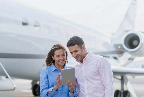 A smiling man and woman, both casually dressed, look at a tablet. They stand in front of a private jet, conveying a sense of business travel.