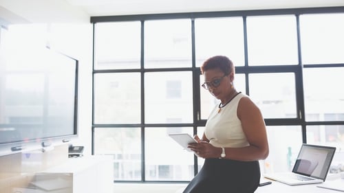 A woman sits on a desk in a bright office, focused on a tablet. Large windows and a laptop suggest a modern and professional work environment.