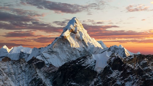 "Snow-covered mountain peak illuminated by golden sunset light, surrounded by darker rocky ridges and a dramatic orange and purple sky"