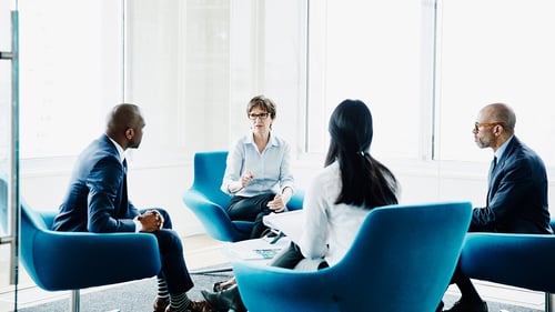 A group of four professionals sits in blue chairs in a modern office with large windows. They are engaged in a serious discussion, fostering a collaborative atmosphere.