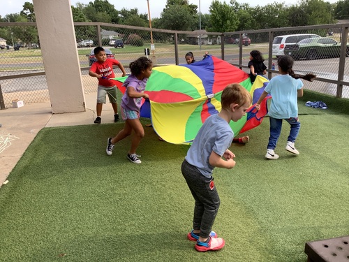 Kids joyfully lifting a bright, multi-colored parachute during group playtime.
