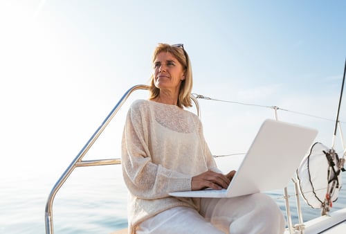 Woman in casual attire works on a laptop aboard a sailboat. She gazes into the distance, conveying relaxation and focus under a clear blue sky.