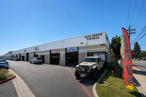 Exterior view of Auto Repair Express, an automotive service shop located in a light gray commercial building with the address number 17242 displayed on the corner. Open service bays feature signs reading “Oil Change” and “Brake Check.” A beige Jeep Gladiator pickup truck with off-road modifications is parked in front of the building. A tall red promotional flag stands along the curb on the right side. The scene is brightly lit under a clear blue sky, with trees, utility poles, and power lines visible in the background.