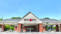A photo of a Bryant Bank branch. The structure features a large peaked roof with the bank’s logo, supported by red brick pillars, and multiple lanes for vehicle service. Green trees are visible in the background.