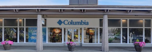 Photograph of Columbia Sportswear storefront with blue and white logo sign above the entrance. The large glass windows display mannequins dressed in outdoor clothing, and the store's interior is visible. The suite number 498 is shown above the door.