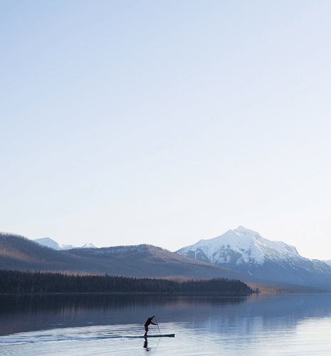 A person in a red outfit paddleboards on a calm lake, with snow-capped mountains and clear blue skies in the background.