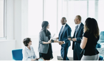 A diverse group of five professionals in business attire engage in discussion and handshake in a bright office, conveying a collaborative atmosphere.