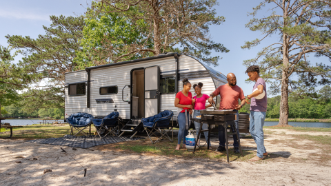 family around a grill at a campground with their camper behind them