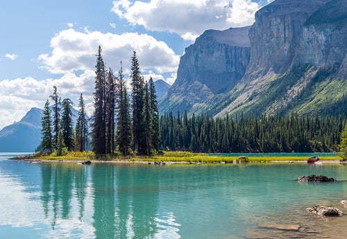 A serene lake scene with turquoise water reflecting tall evergreen trees and rocky mountains. Fluffy clouds scatter across the blue sky, evoking tranquility.