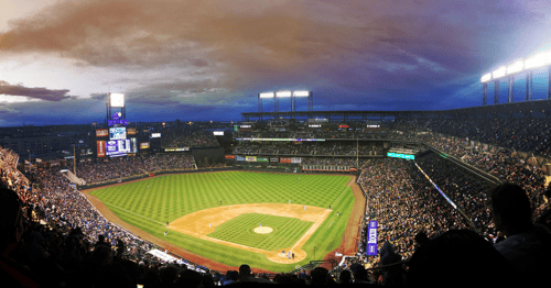 A panoramic view of a packed baseball stadium at dusk, with a vibrant green field under bright lights, and a dramatic, cloudy sky enhancing the atmosphere.