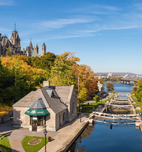 Historic stone building by a canal with locks, surrounded by vibrant autumn trees; distant city and river views under a clear blue sky.