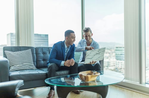 Two men in a modern office sit on a black couch, smiling and discussing content on a laptop. Large windows show a cityscape background.