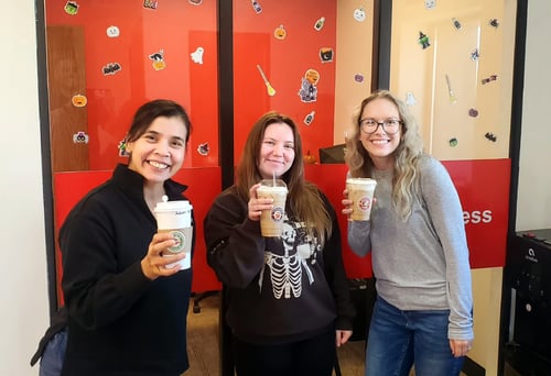 Three women holding coffees in front of a glass window and red wall