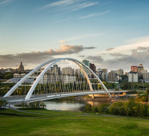 Panoramic view of Edmonton's downtown skyline featuring the iconic white Walterdale Bridge across the North Saskatchewan River, with green parkland in the foreground and golden-hour lighting