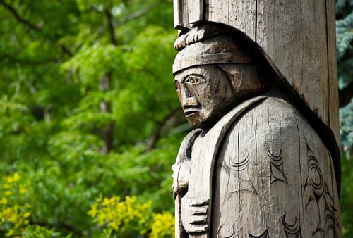 Wooden totem pole carving of a person, featuring intricate designs and a stoic expression. Lush green trees form the serene background.