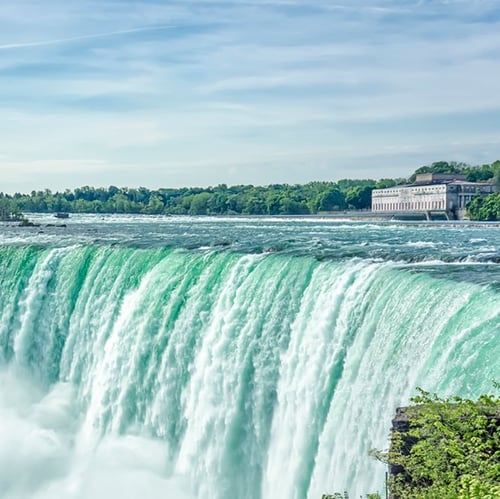 A wide, powerful waterfall with vibrant turquoise water cascades over a cliff, surrounded by lush greenery under a clear blue sky.