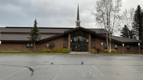 Front view of the Baxter church building showing the south entrance with large glass doors and tall windows beneath a peaked roofline. A steeple rises above the center, with trees, landscaping, and parking spaces visible in the foreground under an overcast sky.
