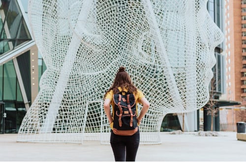 A person wearing a backpack stands facing a large wire sculpture of a head. The figure and modern architecture convey curiosity and exploration.