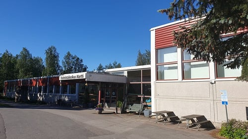 Kuvassa toimintakeskus Martti ulkoa. Matalassa rakennuksessa on harmaa alaosa ja punainen yläosa. Taivas on sininen ja rakennuksen ympärillä näky puita.
