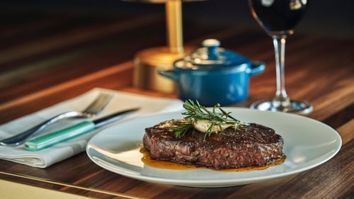 A perfectly cooked steak topped with garlic and rosemary on a white plate, served alongside a glass of red wine and a small blue pot on a wooden table.