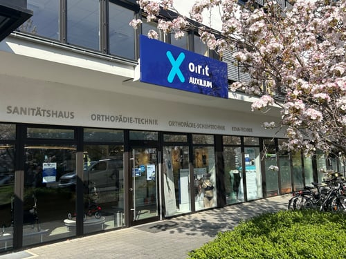 Bild der Filiale Frontal, leicht schräg. Zu sehen ist das Schaufenster sowie ein Teil eines blühenden Kirschbaums rechts oben im Bild.