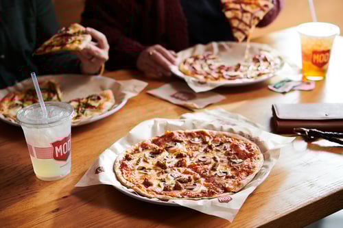 A table at a MOD Pizza restaurant with 3 different individual pizzas and some drinks. 2 people are eating their pizzas.