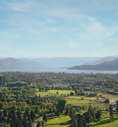 Aerial view of a lush green landscape with a golf course and trees, a cityscape in the middle, and a serene blue lake with distant mountains under a clear sky.