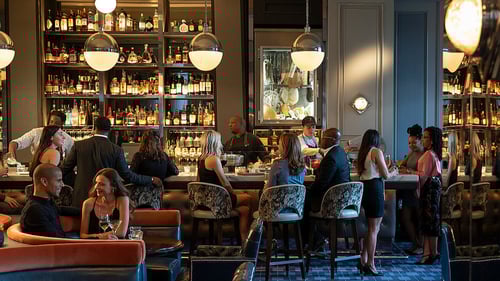 Elegant bar with guests socializing, bartenders preparing drinks, plush seating and shelves stocked with liquor bottles under warm lighting