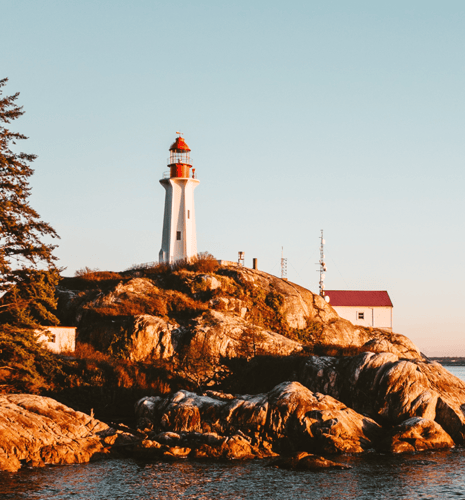 Lighthouse on rocky hill during sunset with warm, golden light. A small white building and trees are nearby, creating a serene coastal scene.