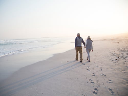 An elderly couple walks hand in hand along a serene beach at sunrise, casting elongated shadows on the sand. Waves gently lap the shore, creating a peaceful atmosphere.