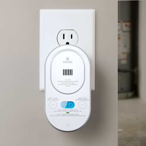 A carbon monoxide alarm plugged into the wall with the display illuminated in blue that shows "CLR" for clear.