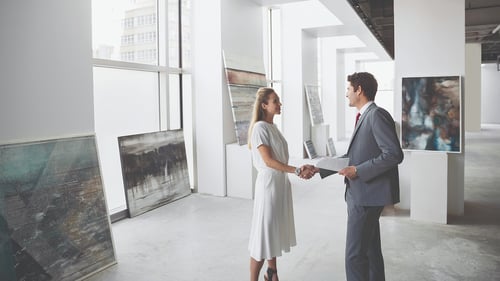A formal handshake between two individuals in an art gallery, surrounded by modern abstract paintings and bright natural light.